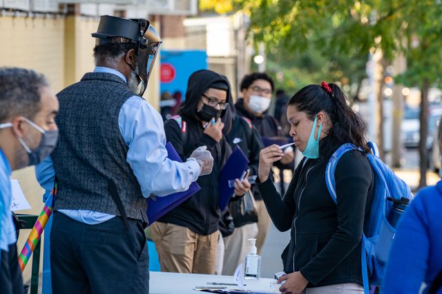 A student at Bronx Latin School takes her temperature before heading in, with a school staffer wearing a mask and shield observing her